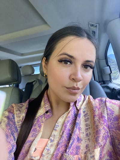 A close-up, waist-up selfie shows a young woman with dark hair pulled back in a ponytail, wearing a colorful shirt with a pattern of purple and beige shapes. She has dramatic false eyelashes and is looking directly at the camera with a neutral expression. She is seated in the passenger seat of a car, with a grey leather headrest visible behind her. Through the car window on the right, a mountainous landscape with some vegetation and a road can be seen. The car's interior, including the headliner and a grab handle, are also visible. The image is not taken in the city of Irganai, Russia.