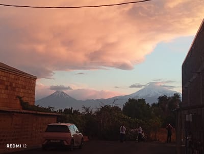 The image depicts a scene in the city of Vedi, Armenia, during what appears to be sunset or sunrise. The sky is dominated by dramatic, fluffy clouds tinged with pink and orange hues, contrasting with a soft blue sky. In the background, two majestic, snow-capped mountains rise above a line of dense green trees. The larger, left mountain has a more defined conical shape, while the right mountain is broader with rugged terrain.

In the foreground, to the left, a building constructed with reddish-brown bricks is partially visible, featuring a dark-tiled roof. In front of this building, a modern, light-colored SUV is parked. The vehicle's license plate, with Armenian characters and numbers, is partially visible.

To the right of the SUV, a dark asphalt surface extends, suggesting a road or parking area. Further to the right, a black metal fence encloses a property. A few individuals are present in the scene. One person stands by the fence, holding a phone up, possibly taking a photograph. Two other individuals, one wearing a white shirt and the other a dark top, are also visible in the middle ground, standing near the fence. The overall atmosphere is serene, capturing a moment of natural beauty with urban elements. The bottom left corner of the image displays the text "REDMI 12."