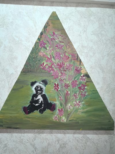 The image is a painting on a triangular canvas that depicts a stylized panda bear sitting in a grassy area next to a bush with pink flowers. The panda is black and white with dark eyes and ears, wearing a white head covering. The background is a blend of greens, pinks, and browns, suggesting a garden or natural setting. The painting is mounted on a textured white wall. There is no identifiable information within the image or its context that would allow for its location to be determined in Taldykorgan, Kazakhstan.