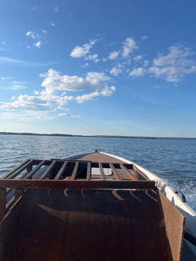 The image captures a scenic view from a boat sailing on a body of water, likely a lake or a wide river, given the distant treeline visible on the horizon. The perspective is from the bow of the boat, looking forward and to the side.
The sky is a vibrant blue, dotted with fluffy white cumulus clouds scattered across the frame, suggesting fair weather. The sun appears to be positioned to the right, casting shadows on the boat's structure and illuminating the water. The time of day seems to be late morning or early afternoon, judging by the brightness of the sky and the angle of the sunlight.
The water is a deep blue, rippling with waves created by the boat's movement. The boat itself is partially visible, with a weathered, rusty metal structure in the foreground. This structure appears to be a part of the boat's equipment, possibly a loading mechanism or a railing. The hull of the boat is white and streamlined, cutting through the water, leaving a visible wake.
The distant shoreline is lined with a dense green forest, indicating a natural, possibly rural or undeveloped, setting. No people are visible in the image, adding to the sense of peaceful solitude. The overall mood is one of tranquility and the enjoyment of a boat ride on a beautiful day. The location is identified as Siukterka, Russia. There is no visible text in the image.