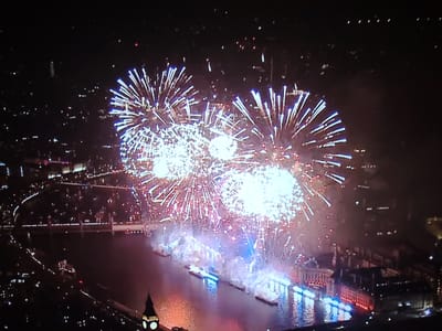 This is an aerial photograph of a fireworks display over a river at night. The fireworks are bright white and blue, bursting in the sky and reflecting on the water below. Several boats are on the river, illuminated by the fireworks. In the foreground, a clock tower, likely part of a historic building, is visible. The background shows the lights of a city, suggesting an urban environment. The location is identified as the River Thames in London, United Kingdom, with the Houses of Parliament and Big Ben visible.