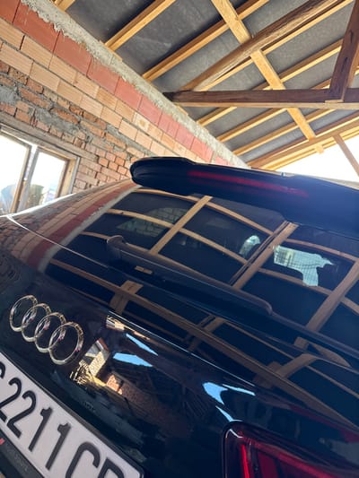 The image displays the rear section of a dark-colored Audi vehicle, likely black, positioned inside a structural enclosure in Paisievo, Bulgaria. The car's trunk lid features the prominent chrome Audi four-ring logo and a black rear windshield wiper. A black spoiler with an integrated red LED light strip is visible at the top edge of the rear window. A partial view of a Bulgarian license plate is present at the lower left, displaying the characters "S 2211 CB" and "Audi Sport" text. Reflections on the vehicle's glossy surface reveal portions of the surrounding interior. The background includes a reddish-orange brick wall with visible mortar joints on the left, featuring a window with wooden framing. Overhead, a ceiling structure is visible, composed of wooden beams arranged in a grid pattern, supporting a dark grey or black material. Natural light illuminates the scene, creating reflections on the car's surface. No individuals are present, and no actions are depicted.