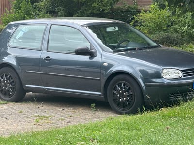 A dark gray Volkswagen Golf IV hatchback is parked on a paved driveway, partially obscured by overgrown grass and foliage. The car is viewed from a side-angle, showcasing its front right wheel and fender, as well as the driver's side door and rear window. A blue and white sticker is visible on the rear window, partially covering the interior. The background consists of dense green trees and vegetation, suggesting a natural or suburban environment. There are no people or any visible actions taking place. The car appears to be stationary. The setting is consistent with a residential area in Bemmel, Netherlands.