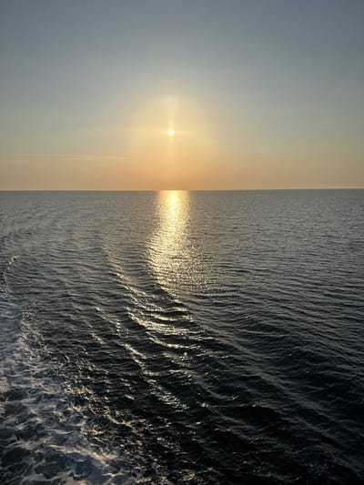 The image depicts an open expanse of water under a sky at either sunrise or sunset, observed from a moving vessel off the coast of Kato Karioutes, Greece. The horizon line is clearly defined and straight. The upper third of the frame shows a sky transitioning from a light blue at the top to a warm orange-yellow hue near the horizon. The sun, a bright circular point, is positioned slightly left of the image's vertical center, low on the horizon. A prominent vertical column of sunlight reflects intensely on the water surface, extending from the sun towards the foreground, creating a shimmering golden path. The remaining water surface is a dark blue-grey, displaying numerous small, uniform waves. In the lower left quadrant, a distinct white, turbulent wake from a moving vessel is visible, contrasting sharply with the darker sea. No landmasses, structures, individuals, or other watercraft are present in the scene.