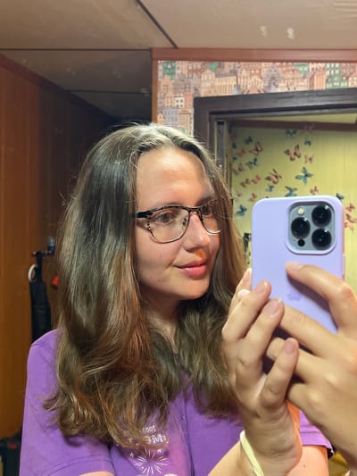 The image is a selfie taken by a young woman in a vertical orientation. She is wearing a purple t-shirt and has long, wavy brown hair. She is also wearing glasses with thin, dark frames. Her eyes are visible, and her expression is neutral. The woman is holding a light purple phone in her right hand, and her left hand is also visible, resting on the phone. The phone has a triple-lens camera system.
The background of the image is a mirror reflecting a room. The wall behind her appears to be wallpapered with a pattern of small buildings and butterflies. A wooden frame is visible, suggesting the mirror is part of a larger piece of furniture or decoration. The lighting suggests it is daytime, possibly indoors. There is no discernible weather information. The overall impression is of a casual, personal moment captured by the subject herself. There is no visible text in the image.