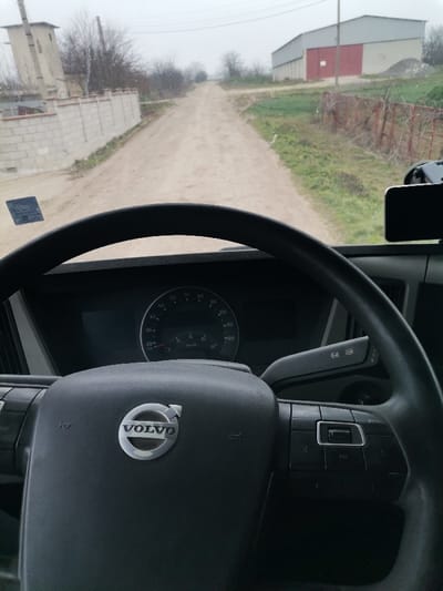 The image presents a first-person view from the driver's seat of a Volvo vehicle, situated on an unpaved dirt road in Kardam, Bulgaria. The black Volvo steering wheel, bearing the silver Volvo logo, dominates the lower foreground, with dashboard instruments including a speedometer and tachometer visible behind it. A smartphone is mounted on the right side of the dashboard. A rectangular sticker is affixed to the lower-left of the windshield, which shows minor smudges.
The dirt road, marked by tire tracks, extends centrally into the distance under an overcast sky. To the left, a low concrete block wall parallels the road, behind which stands a tall, light-colored concrete utility structure and sparse, bare trees. To the right, green vegetation and a rusty metal fence border the road. In the mid-ground right, a large, light-colored industrial-style building with a distinct red section is visible, fronted by a pile of aggregate and a utility pole.