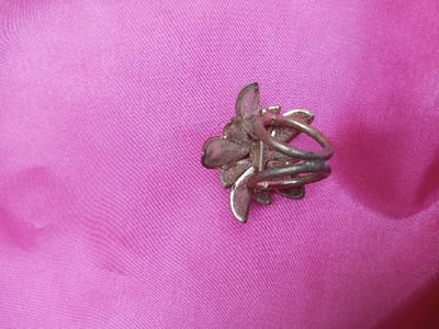 The scene depicts a metallic ring resting on a vibrant pink fabric surface. The location is Qarabucaq qəsəbə, Azerbaijan. The ring is oriented on its side, showcasing its double-banded shank and the underside of its elaborate, flower-like decorative head. The head is constructed from multiple layered elements of what appears to be silver-toned metallic mesh or filigree, exhibiting a somewhat tarnished or aged finish. The underlying fabric is a finely woven, brightly colored pink material, likely satin or a similar textile, with visible diagonal weave patterns and slight creasing, particularly in the lower right quadrant. No individuals are present, and no actions or interactions are occurring. The environment consists solely of the fabric surface and the ring, under what appears to be ambient indoor lighting.