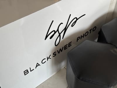 The image shows a white wall with black lettering. The lettering reads "bsp" in a stylized script font, and "BLACKSWEE PHOTO" in a sans-serif font. Two black hexagonal stools are partially visible in the foreground. The setting appears to be an indoor space, possibly a studio or a waiting area. There is no visible indication of the location in Kedawung, Indonesia.