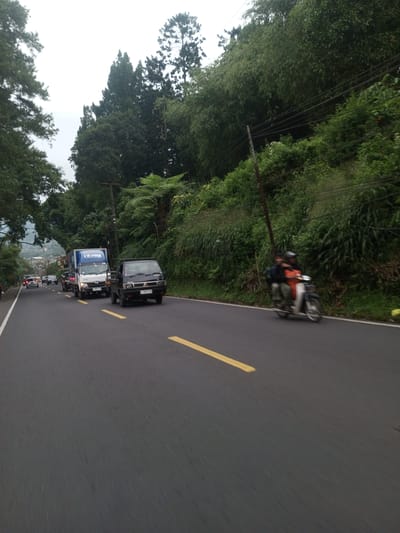 The image depicts a road scene with multiple vehicles and a motorcycle traveling along an asphalt road marked with yellow dividing lines. A blue and white truck is followed by a small black pickup truck, along with other unidentified vehicles further in the distance. A motorcycle with two passengers is traveling on the right side of the road. The road is bordered by dense vegetation on the right and trees on the left, with utility poles and overhead wires visible above the foliage. The overcast sky suggests daytime. The location of Taunggyi, Myanmar, is not verifiable based on the information provided.