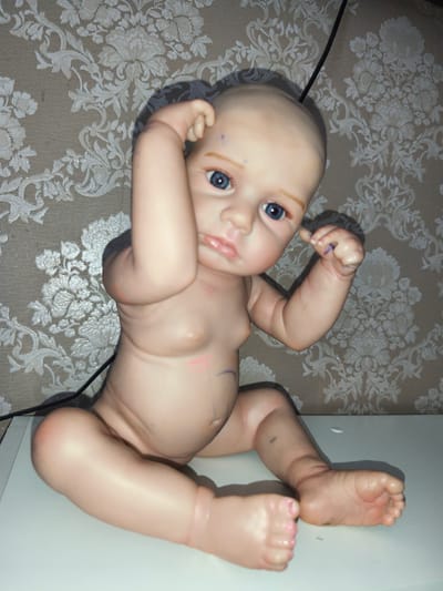 This media file features a doll that resembles a baby. The doll has blue eyes, light-colored eyebrows, and a bald head. It is positioned in a seated posture, with its legs bent and arms raised near its head. The doll's skin tone is light, and there are some faint markings or smudges on its torso and legs. It is sitting on a flat, white surface, possibly a table or shelf.
The background consists of wallpaper with a repeating floral pattern in white against a beige or light brown textured background. There is a black cable or cord visible in the upper right corner of the image, trailing behind the doll.
The overall lighting suggests an indoor setting, and the time of day cannot be determined. There is no discernible weather or specific location cues beyond the wallpaper pattern. The doll's pose and facial expression do not convey any specific emotion, appearing neutral or slightly curious. No text is visible in the image. The context of Rahachow, Belarus, is provided externally and is not evident from the visual content itself.