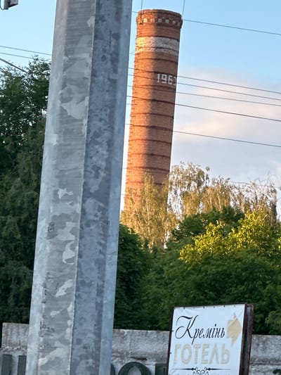 The image depicts an outdoor scene in the city of Kremenets, Ukraine. A tall, brick smokestack, possibly dated 1963 as indicated by markings on its surface, is visible against a partly cloudy sky. In the foreground, a large, galvanized metal pole, likely a utility pole or structural support, stands prominently. Lush green trees and foliage surround the area. A sign with the text "КРЕМІНЬ ГОТЕЛЬ" (Kremen Hotel) is positioned near the bottom right of the frame, suggesting the presence of a hotel. The overall environment appears to be an industrial or semi-industrial area, possibly on the outskirts of the city.