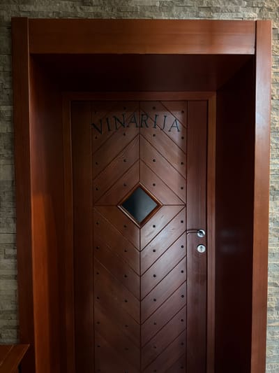 The image shows a dark wooden cabinet in an interior setting. The cabinet's door features a diamond-shaped window in the center and is decorated with parallel grooves and small, round dark metal accents. The word "VINARIJA" is vertically inscribed on the left side of the door. A key is inserted into one of the locks on the top center of the cabinet. The cabinet is positioned against a wall covered with light gray stone-like tiles. Based on the word "VINARIJA", which translates to "Winery," it is highly probable that this object exists at the Vinarija, or Winery, in Krašići, Montenegro.