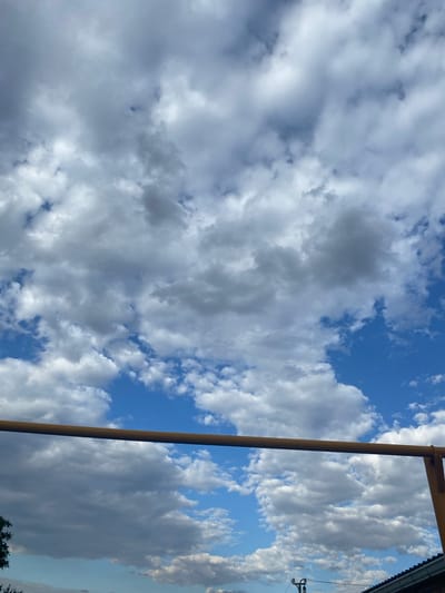 The image captures a bright, overcast sky filled with dramatic, textured cumulus clouds. A thin, rusty metal bar, possibly part of an outdoor structure or play equipment, extends horizontally across the lower portion of the frame, obscuring a sliver of the sky. Below the bar, the edge of a dark, tiled roof is visible on the right side of the image, hinting at a residential or urban setting. A small portion of a green tree appears on the far left. The sky itself is a vibrant blue peeking through the white and grey clouds, suggesting a day with variable weather conditions. While the location is specified as Taganrogsky, Russia, the visual information within the frame does not offer specific landmarks or contextual clues to confirm this geographically.