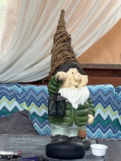 A gnome garden statue stands in front of a blue and white chevron pattern blanket. The gnome is wearing a green jacket, a black hat, and has a white beard. It is holding a black lantern in its right hand. The gnome's hat is conical and made of twigs. Behind the blanket, there is a white curtain. In the foreground, there is a gray table with several objects on it, including a small white container and what appears to be a dark case. The scene is outdoors, possibly on a patio or deck. The image does not contain any explicit location information for Bersenevka, Russia.