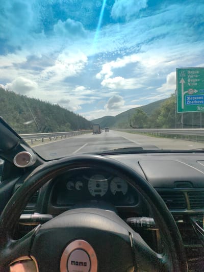 The image, taken from the driver's perspective inside a car, shows a highway scene. The sky is bright blue with scattered clouds, and sunlight is casting a lens flare from the top right. The road ahead is a multi-lane highway with a guardrail on the left and a forested hillside on the right. A truck is visible in the distance on the highway. A green road sign is prominently displayed on the right, indicating directions to Varna, Vidin, Zherkovo, and a distance of 500 km. The interior of the car shows a dashboard with prominent gauges, a steering wheel with a "Momo" logo, and a turn signal lever. The location is identified as being near the city of Potop, Bulgaria.