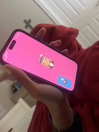 A person is holding an iPhone showing a TikTok video call. The main video features an avatar of a person wearing a traditional Arab headdress, while the smaller inset video shows an avatar of a female-presenting person. The phone is held in front of a person wearing a red fleece hoodie. A gold beaded bracelet is visible on the person's wrist. The background includes a white door with recessed panels, a green four-pointed object resembling a stylized plus sign, and a white dresser or cabinet. The time on the phone is 00:14, and the cellular connection displays 5G. The location cannot be determined from this image.