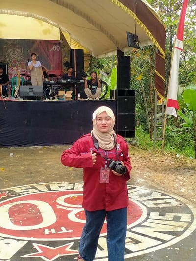 A young adult female stands in the foreground, facing the camera with a neutral expression. She wears a beige hijab adorned with a small white flower on the left, a red long-sleeved collared shirt, and blue jeans. A black camera strap is around her neck, and she holds a black DSLR camera with a lens in her left hand. A red identification badge hangs from a lanyard on her right side, partially visible, with "JOVA PRATAMA" printed on her shirt. She has her left thumb hooked near her shirt pocket. She stands on a paved surface featuring a large circular red and white logo, which includes the number "80" and stylized text.
In the midground, a temporary outdoor stage is set up under a cream-colored tent canopy with a dark brown and gold valance. A large banner on the stage backdrop features a lion's head logo, text partially reading "PONPES SENJARA...", and the number "80." On the stage, a female performer in a light-colored dress stands at a microphone on the left, singing with her right hand raised. To her right, a male keyboardist is seated. Further right, a male videographer in a dark t-shirt and light pants squats, holding a video camera and smiling at the viewer. A bicycle is partially visible behind him. Black speaker stacks are positioned on both sides of the stage.
The background consists of dense green foliage, trees, and banana plants, indicating an outdoor, natural environment. To the right of the stage, a tall white and red Indonesian flag flies vertically on a pole. The scene takes place in the city of Tamansari, Indonesia.