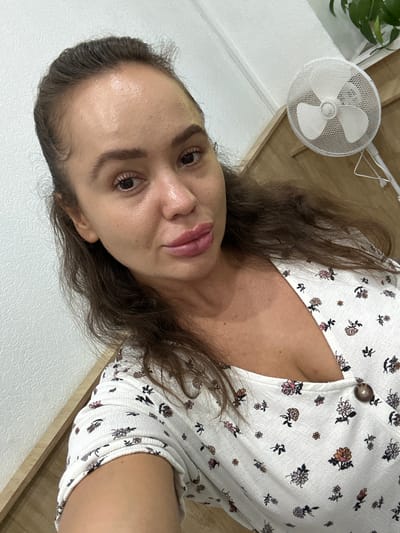 A close-up, high-angle selfie shows a young adult female with brown hair pulled back and loose strands framing her face. She is wearing a white V-neck top with a floral pattern and a single brown button. Her lips are full and appear to be coated in lip gloss. Her skin is visible, showing some texture and minimal makeup. Behind her, a white oscillating fan is positioned on a wooden surface, and a portion of a green plant is visible in the upper right corner. The background consists of a white textured wall and a wooden surface. The image appears to be taken indoors, likely in a residential setting. There is no visual information present in the image that would indicate the location is Duvankul', Russia.