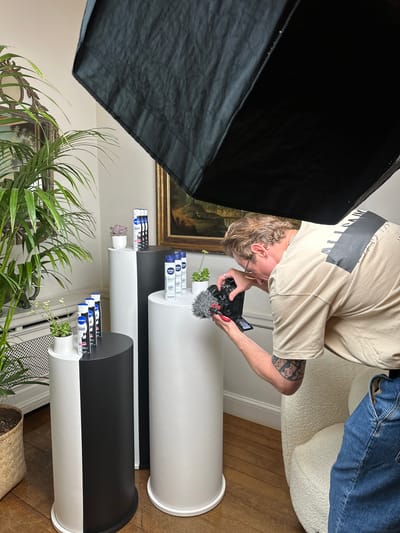 In a room with wooden flooring, a person wearing a beige t-shirt and denim jeans is filming a product display with a digital camera. The products are aerosol cans, likely deodorants, arranged on cylindrical pedestals of varying heights and colors, predominantly white and black. One pedestal is split vertically between black and white. The room contains a large potted plant with green foliage to the left, a framed artwork above a radiator on the wall, and a textured armchair to the right. A large, black softbox light is positioned overhead, casting light onto the scene. The overall impression is a studio or a well-decorated room set up for a product photoshoot or video recording. The location is the city of Chichester, United Kingdom.