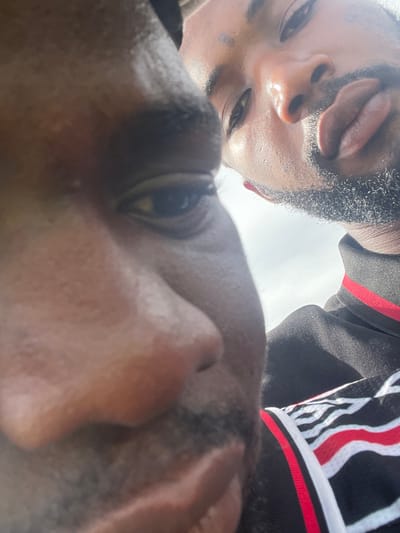 This close-up, low-angle photograph, taken in Ogbogbo, Nigeria, captures two individuals looking down towards the camera. The perspective is from below, giving an intimate, upward gaze. In the foreground, the right side of one person's face, including an eye and part of the nose, is visible, occupying the lower right. Their skin is dark, and their expression is not fully discernible.
Above and behind this individual, another person's face appears upside down, looking directly into the camera. This individual has dark skin, short dark hair, and a beard, with a neutral to slightly smiling expression. They are wearing a dark top, possibly athletic wear, featuring distinctive red and white striped patterns on the collar or shoulder.
The scene is intensely bright, with a large, almost completely white background suggesting an overcast sky or strong overhead light during the daytime. This strong backlighting illuminates the subjects' faces, creating highlights and a soft glow around their outlines. The overall impression is a casual, direct, and brightly lit shared moment. No text is visible in the image.