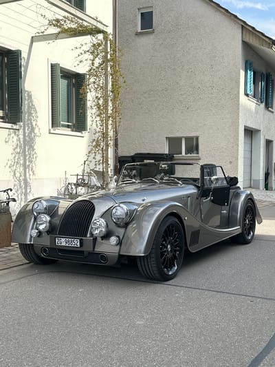 The image shows a silver Morgan sports car parked on a street. The license plate reads "ZG-98852". The car is parked alongside a building with light-colored stucco and windows with green shutters. The street is paved with asphalt. A vine covers part of the building in the background. The location is inferred to be Rafz, Switzerland, based on the license plate code (ZG), which corresponds to the canton of Zug in Switzerland.