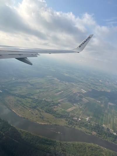 An aerial view from a Ryanair airplane shows the wing and flaps over rural Zakroczym, Poland. The wing, marked with "RYANAIR," is set against a backdrop of scattered clouds and blue sky. Below, a landscape of agricultural fields and forests stretches to the horizon. A river meanders through the terrain, bordered by dense woodland. The fields show grid-like patterns and are interspersed with small villages and roads. The overall visibility is slightly obscured by haze.