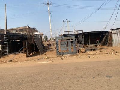 This image presents a wide view of an outdoor metal fabrication or scrap yard situated along a paved road in Kaduna South, Nigeria. The sky is clear and bright, indicating daylight conditions.
In the foreground, a dark asphalt road surface is visible, bordered by a dusty, unpaved shoulder scattered with various debris, including small pieces of metal and litter.
On the left, an open-fronted, multi-level structure serves as a workshop or storage unit. It is constructed from metal framing and corrugated sheets, heavily stacked with bundles of dark metal rods, square tubing, and flat metal sheets. A silver-colored metal ladder leans vertically against the stacked inventory on the left side. The upper level of this structure also holds metal components.
Toward the center and right, two additional open-fronted workshops are visible. These structures consist of corrugated metal roofs and partial walls, supported by wooden poles and metal frames. Sections of these structures, particularly the wooden elements and corrugated sheeting, display significant darkening consistent with charring or heavy use.
Several large, unfinished or raw metal gate frames and grates, primarily dark grey and blue, are arranged on the dirt ground between the paved road and the workshops. A prominent blue metal gate frame, approximately 1.5 meters in height, stands centrally.
Three individuals are present within the scene. In the central-right workshop, a person wearing a dark red shirt and dark trousers is bent over, actively handling or working on metal pieces near a stack of materials. To their left, another individual in dark attire stands upright near a metal structure, engaged in a task involving metal. A third individual, also in dark clothing, is partially discernible further back and to the left, adjacent to the stacked metal inventory.
Overhead, multiple concrete utility poles with numerous interwoven power lines traverse the frame from left to right. A dark-colored motorcycle is partially visible behind a stack of metal components in the central area, towards the back of the rightmost workshop. The ground within the workshop areas is unpaved earth, uneven, and covered with metal scraps and waste.