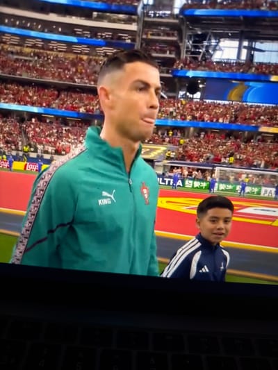 This media file captures a close-up view of a young boy standing next to a man, both appearing to be on a football pitch surrounded by a large crowd. The man, prominently featured in the foreground, is looking upwards and to the side, his expression is somewhat unclear due to the angle and image quality. He is wearing a teal-colored jacket with a red emblem on the chest and a "KING" logo on the sleeve. The boy, positioned behind and to the right of the man, is wearing a blue and white Adidas tracksuit. The background reveals a stadium filled with spectators dressed in red, suggesting a football match or a related event. Banners and stadium structures are visible, including what appear to be large blue vertical displays. The scene is brightly lit, indicative of daylight or well-lit stadium conditions. Visible text includes "Lay's", "KING", and some Chinese characters on a banner. The overall context suggests a sporting event, possibly a match involving the man or a ceremony associated with it.