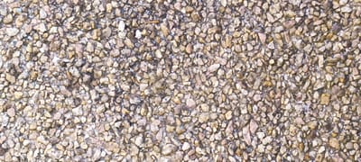 The image shows an aerial view of a ground surface composed of small, irregularly shaped stones and pebbles. The stones are a mix of earthy tones, including beige, brown, and gray, with occasional flecks of white and darker gray. The texture of the ground is rough and uneven due to the size and arrangement of the pebbles. Some larger stones are visible interspersed among the smaller ones, creating a varied surface. The lighting suggests it is daytime, with shadows cast by the stones indicating a natural light source. There are no visible people, objects, or text in the image. The scene depicts a natural or man-made ground covering, possibly a path, driveway, or an exposed aggregate surface. The context of Winnenden, Germany, does not provide specific visual cues within the image itself, but it establishes a geographical location. The image is a close-up, texture-focused shot, emphasizing the materiality of the ground.