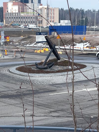 The image displays an urban roundabout in Espoo, Finland, featuring a prominent anchor sculpture as its central monument. A large, dark-colored, weathered anchor with a distinct wooden stock is positioned vertically within the roundabout's central island. This island consists of dark earth and rocks, encircled by a ring of light-colored cobblestone paving. The surrounding roadway is asphalt.

In the mid-ground, a white cargo van and other vehicles are observed on a road beyond the roundabout. Extensive multi-story building construction, including incomplete facades and a visible crane, is underway in the background. A large white information sign, partially obscured, displays Finnish text, including the word "Espoo," likely indicating the construction project. Multiple yellow triangular warning signs, depicting a pedestrian, are present, alongside a red circular sign indicating a 3.4-meter height restriction. Yellow and white traffic barriers line sections of the road, and a long blue corrugated structure stands in the mid-ground to the right. Bare tree branches with visible buds occupy the immediate foreground, slightly blurring the lower portion of the scene. Patches of dirty snow are scattered on undeveloped ground beside the roads. A distant treeline, comprising bare deciduous and some evergreen trees, is visible beneath an overcast sky. No individuals are discernibly present within the captured scene.