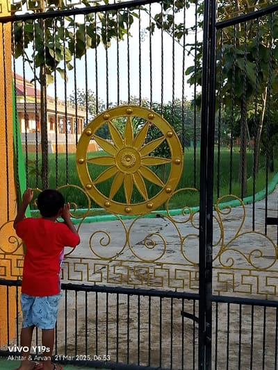 A young boy, wearing a red shirt and blue-and-white patterned shorts, stands behind a black metal gate with ornate gold designs. The gate has a decorative gold wheel-shaped design on the left side. The gate is open and the boy is looking through it. The ground is covered in green grass. The scene is located in Kecamatan Anjatan, Indonesia, at latitude: -6.417 and longitude: 107.978. The background is a lush green field. A building with a yellow-painted wall can be seen through the gate.