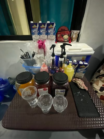 The image depicts a setup for serving drinks, likely in a commercial or event setting, in San Jose del Monte, Philippines. The primary objects are drink dispensers, containers of syrup or flavoring, milk cartons, plastic cups, and assorted preparation items.

In the background, a white cooler with a black spigot is visible. To its right are several blue and white cartons of "Full Cream Milk." Next to the milk cartons is a yellow squeeze bottle, presumably containing a liquid ingredient. Behind these, a pink cloth or towel is draped over a dark brown textured surface, which appears to be a tabletop or serving counter. This surface has a black perforated mat embedded in it.

In the foreground, several large, clear plastic jars with black lids are arranged. These jars contain colorful liquids, suggesting different flavors of drinks, possibly syrups or pre-mixed beverages. Some of these jars have labels, though the text is mostly illegible. Beside the jars, clear plastic cups with lids are stacked, with some appearing to be filled with liquid. Black straws are neatly organized in a clear plastic container.

A small, decorative red backpack with a cartoon character is positioned near the top left of the frame, adding a whimsical touch to the scene. The overall lighting suggests an indoor setting, possibly with artificial light. There are no people visible in the image. The arrangement of items indicates preparation for a drink service, potentially for a party, gathering, or a stall. The visible text on the milk cartons reads "Full Cream Milk." Some labels on the jars and cups have numbers like "58" and "88".