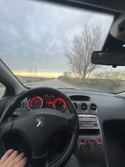 The image shows an interior view of a car, specifically a Peugeot, driving on a rural road. The dashboard and steering wheel are visible, with the Peugeot logo prominent on the steering wheel. The speedometer and RPM gauge have red indicators. Through the windshield, a straight road stretches into the distance, flanked by bare trees and fields. The sky is overcast with a gradient of grey clouds at the top and faint yellow light on the horizon. The location is Ivanski, Bulgaria. A hand is partially visible in the bottom right corner. The car's central console with buttons and vents is also visible.