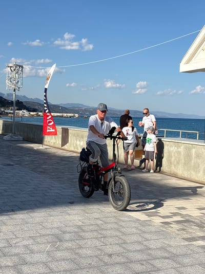 The image depicts a man riding an electric bicycle on a paved promenade by the sea. He is wearing a white t-shirt, grey shorts, and a grey baseball cap. In the background, there are other people walking and standing, as well as the sea and distant mountains. A tall, ornate lamp post is visible to the left, and a flag with a red portion displaying the Cyrillic letters "РОДИНА" is planted in the ground. The scene is illuminated by bright sunlight, casting shadows on the promenade. The location is identified as the Alushta Embankment, Alushta, Crimea, Ukraine.