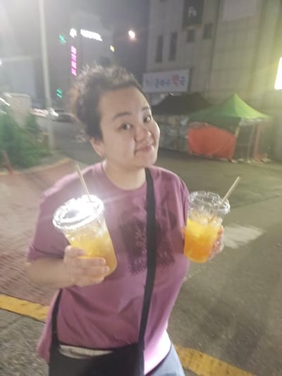 In Eumnae-ri, South Korea, at night, a woman with dark hair pulled up is holding two plastic cups filled with a yellow-orange beverage, likely juice or a mixed drink, each with a straw. She is wearing a pinkish-purple t-shirt and a dark strap across her chest, suggesting a bag or camera strap. The background shows a dimly lit urban environment with buildings and tents, possibly a market or outdoor event. A neon sign with Korean characters is visible on a building to the left. The ground is paved, and a yellow line is present on the left side of the image. The overall lighting is low, with some light sources creating a bright glare in the image.