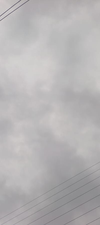 A cloudy sky fills the frame with varying shades of gray. Wisps and patches of lighter and darker tones create an indistinct texture. The upper left and lower right corners are bisected by dark, linear power lines. The lines converge toward the bottom, creating a stark contrast with the soft, diffused sky.