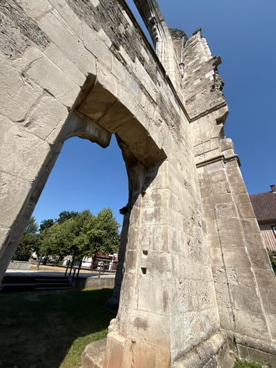 The image shows a close-up, low-angle shot of the ruins of the Walkenried Abbey in Walkenried, Germany. The dominant feature is a large stone archway and a portion of a weathered stone wall, made of large, rectangular blocks. The stonework shows signs of age and erosion, with some rough textures and discolored patches.

Through the archway, a clear blue sky is visible, along with a section of a grassy area with some trees and a paved pathway. Beyond the trees, a building with a brown tiled roof and light-colored walls can be seen. The sunlight casts shadows on the ruins and the ground, indicating a bright, sunny day. There are no people or animals visible in the image. The ruins appear to be part of a historical structure, likely an abbey given the location.
