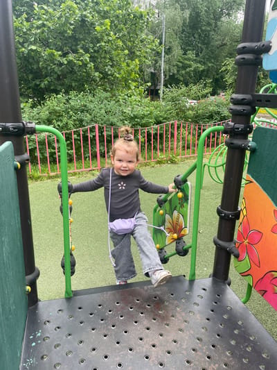 The image depicts a young girl, approximately four years old, standing on a metal platform at a playground. The girl is wearing a purple shirt with a white star, blue jeans, and a light purple purse. She is holding onto a green ladder-like structure. She is facing the camera with a slight smile. The platform is made of a black metal with a pattern of holes. The background of the image shows a pink metal fence and lush greenery. The location is a playground in Moscow, Russia, with the coordinates 55.659, 37.591.