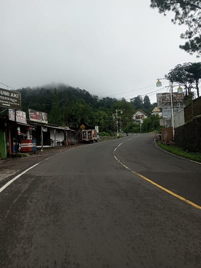 This image depicts a winding asphalt road through a mountainous, semi-urban landscape in what is described as the city of Bangtsho, Bhutan, under an overcast sky. The road, appearing damp, features a solid white line marking its left edge and a yellow line delineating the center for a gentle right curve.
On the left side of the road, a series of low-rise commercial structures or stalls are present. A prominent sign above one reads "BUMI AKI Heritage" with the additional text "Autentik Jawa Barat." Below this, a red "BENSIN MINI" fuel dispenser is visible. Further along this side, a small white utility truck, possibly a mobile vendor, is parked off the road. The background on the left features densely forested mountains partially obscured by mist or low clouds.
The right side of the road is bordered by a stone retaining wall, partially covered with greenery. Further up, a large, billboard-like sign structure, possibly related to "BUMI AKI," stands alongside the road, accompanied by poles from which decorative, lantern-like objects are suspended. Beyond the wall and sign, several distinct houses with light-colored facades and dark roofs are visible amidst trees, one featuring a small dome or turret. A single motorcycle is seen traveling uphill on the road in the distance. Overhanging pine branches are visible in the upper right corner of the frame. No pedestrians are discernibly present.