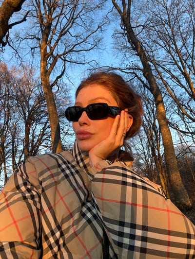 The image captures a single individual, appearing female, outdoors in a wooded environment. The subject, positioned centrally, is illuminated by warm, low-angle sunlight, indicative of either sunrise or sunset, casting a golden hue on their skin and the surrounding bare deciduous trees. The individual wears black, oval-shaped sunglasses with a discernible 'PRADA' logo on the left temple (viewer's right) and a beige puffer jacket featuring a prominent black, white, and red plaid pattern. Their right hand (viewer's left), adorned with a thin ring on the ring finger, is casually resting against the jawline. The background consists of numerous leafless trees against a clear blue sky. While specified as being in the city of Buriani, Georgia, the image does not contain specific visual landmarks to confirm this precise geographical location.