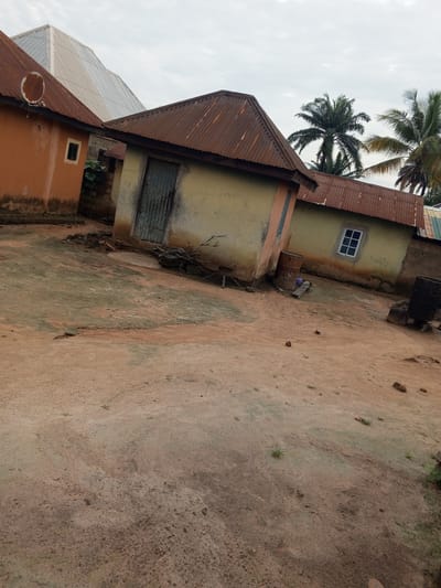 The image shows an outdoor scene in a rural setting, likely Gindin Dutse, Nigeria. The focus is on several small, weathered buildings with corrugated metal roofs. The primary building in the foreground is painted a pale yellow and has a rusty metal door and a small, white-paned window. To its left is another building, painted orange, with a satellite dish on its roof. Behind these, a larger building with a light-colored, angular roof is visible. The ground is dusty and sandy, with scattered rocks and sparse vegetation. Two large metal barrels, one rusty and one black, are present near the buildings, suggesting outdoor storage or utility. Palm trees are visible in the background, a common feature in tropical climates. The sky is overcast, indicating a cloudy day. There is no visible activity or people in the frame, giving a sense of stillness. Some faint, illegible markings or graffiti are present on the wall of the yellow building. The overall impression is of a humble, somewhat aged residential or communal area.