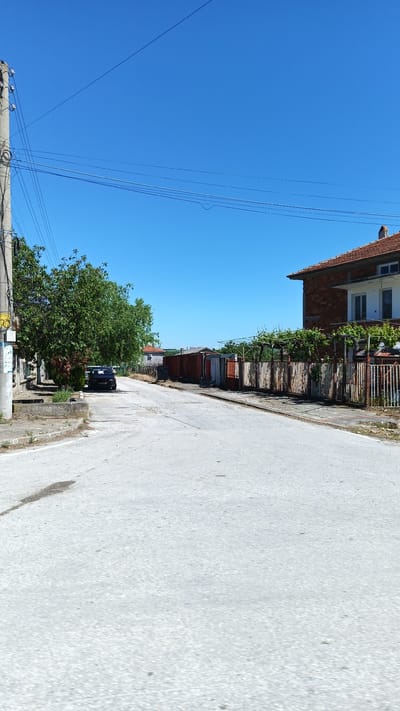 The image depicts a street scene in the village of Seltsi, Bulgaria. A narrow, gray concrete road runs through the frame, showing wear and a few darker spots. On the left, a weathered telephone pole stands with multiple wires extending across the clear blue sky. Adjacent to the pole, a leafy green tree partially obscures a dark-colored sedan parked alongside the road. On the right, a two-story brick house with a red tile roof is visible. A weathered wooden fence runs along the right side of the road, supporting vegetation. The street is mostly empty, lending to the quiet ambiance of the scene.