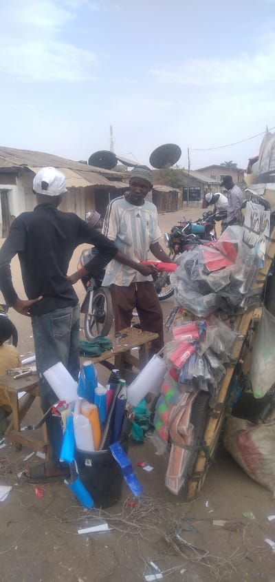 The image depicts a bustling street scene in Chikaji, Nigeria, under an overcast sky. In the foreground, a black bucket is filled with numerous rolls of colorful vinyl or sticker material, predominantly white, blue, and orange. Adjacent to it, a small wooden stool holds a pair of scissors and other cutting tools. The ground is dirt, littered with paper scraps and dried roots.
Two men are central to the midground. On the left, a man wearing a white baseball cap, a black long-sleeved shirt, and blue jeans stands with his hands on his hips, facing right. He observes another man, positioned slightly to the right, who wears a light blue and white striped Adidas Argentina soccer jersey, brown pants, and a patterned cap. This second man holds a red rectangular object in his hands, presenting it towards the first man, suggesting a transaction or display. Behind these men are several parked motorcycles. To the far right, a makeshift wooden stall or cart is laden with various goods, many wrapped in clear plastic, including red and black items.
In the background, a street extends, flanked by single-story buildings with corrugated metal roofs. A sign for "SPORT CENTRE" and "OBELI" is partially visible on one building. Above the rooftops, a tall telecommunication tower and two large, dark-colored satellite dishes are prominently visible against the hazy sky. Other individuals are present further down the street, some interacting with motorcycles, indicating active street life.