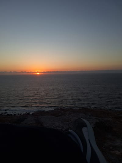 The image captures a serene sunset over the ocean, viewed from a rocky cliffside. The sun, a bright orange orb, is partially submerged below the horizon, casting a warm glow that transitions from vibrant orange near the sun to a soft yellow and then to a muted grey in the upper portion of the sky. The ocean stretches out, a vast expanse of rippling water, reflecting the twilight hues. The water's surface appears textured, with subtle waves and a hint of foam along the distant shoreline.

In the foreground, a person's leg, clad in a dark garment and a black and white sneaker, is visible. The shoe, with its distinctive swoosh logo, suggests casual wear. The person is positioned on the edge of a rocky outcrop, which dominates the right side of the frame. This terrain is rough and uneven, with visible stones and sparse vegetation.

The overall mood is peaceful and contemplative. The fading light and the expansive natural setting evoke a sense of calm and solitude. The time of day is clearly sunset, given the sun's position and the atmospheric colors. The location appears to be a coastal area, with the sea and cliffs being prominent features. There is no visible text in the image.