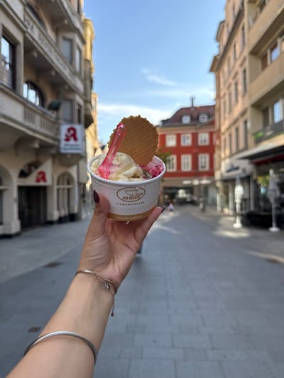 A street scene shows a person holding a cup of gelato with a wafer and a pink plastic spoon. The gelato is from "Ido Gelato Eismanufaktur" and is held out against a backdrop of buildings in a European city, identified as Aschaffenburg, Germany. One sign with a red "A" is visible, indicative of a pharmacy ("Apotheke" in German). The buildings are various colors, with a red building in the middle distance. The street is paved, and other people can be seen walking in the distance. The person's hand and wrist, adorned with a silver bracelet and bangle, are in the foreground. The image appears to be taken from the perspective of the person holding the gelato.