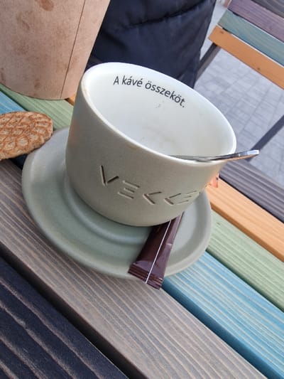 The scene depicts an outdoor setting in Odorheiu Secuiesc, Romania. A largely empty, light green ceramic coffee cup rests on a matching saucer on a multi-colored wooden slatted surface, likely a table or bench. The cup's exterior features the embossed word "VEKKE." Its off-white interior displays the black text "A kávé összeköt." (Hungarian for "Coffee connects.") and shows traces of a light brown liquid residue. A silver metal spoon is positioned inside the cup. On the saucer, a brown, rectangular sugar packet is present. To the left of the cup, a textured, round, light brown cookie or biscuit lies on the wooden surface. In the upper left quadrant, a portion of a tall, light brown disposable paper cup with visible stains is present. Behind these items, a dark, textured fabric surface, consistent with an article of clothing, is partially visible. In the upper right quadrant, a section of a multi-colored wooden chair back is visible, featuring slats in similar shades to the table. The wooden surface is composed of planks painted in various hues including black, dark grey, light grey, blue, green, and brown.