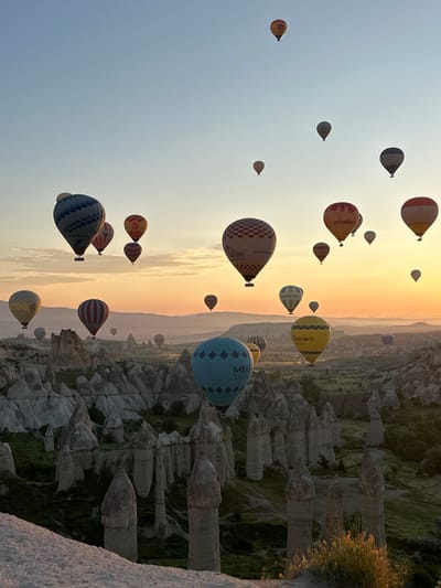 The image captures a breathtaking sunrise scene over the unique geological landscape of Göreme, Cappadocia, Turkey, featuring numerous hot air balloons. The sky transitions from a clear, pale blue at the top left to vibrant golden and orange hues near the horizon line, indicating the early morning light.

Dozens of colorful hot air balloons of various sizes and patterns ascend gracefully across the sky. These balloons display a range of designs, including solid colors, stripes, and checkered patterns. Many also feature visible text and branding. Among these, a large yellow balloon prominently displays "KAPADOKYA BALLOONS". A significant blue balloon carries the text "MEGA TRAVEL", while two separate checkered balloons—one with white and brown squares, another with grey and white—are branded with "Sultan Balloons". An orange-red balloon clearly shows "priceless", and a green and white balloon reads "WOODSTOUR". Another smaller yellow balloon has partially visible text, "Cella".

Below the balloons, the distinctive "fairy chimney" rock formations and valleys, characteristic of Göreme's volcanic tuff landscape, stretch across the terrain. These ancient geological structures are partly illuminated by the soft, emerging sunlight, with patches of green vegetation clinging to their sides. The scene depicts a popular hot air ballooning event, offering panoramic views of this UNESCO World Heritage site during optimal weather conditions, evoking a sense of wonder and tranquility.