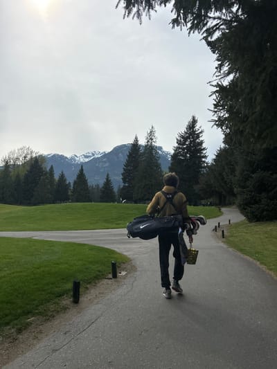A person is walking on a paved path at a golf course, carrying a golf bag. The bag is a dark color and has a Nike logo on it. The person is wearing a tan jacket and dark pants. They are also carrying a small rectangular object, possibly a scorecard.

The path runs alongside a grassy area. The background includes evergreen trees and a mountain range with snow on the peaks. The sky is overcast.

The location is Squamish, Canada, likely at the Squamish Valley Golf Club.