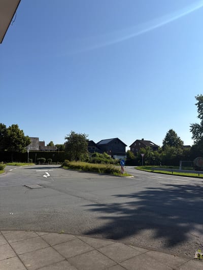 This is a forward-facing, eye-level, outdoor photograph of a street intersection in the city of Beckum, Germany. The sky is clear and blue, with a slight lens flare in the upper right quadrant. The foreground consists of a paved sidewalk with square concrete tiles, meeting a two-lane asphalt road. The road curves to the left and right, with a circular island in the center of the intersection.
On the circular island, there is a sign with a blue arrow indicating a mandatory right turn, and a red and white striped pole. The road is marked with white lane lines and a white arrow indicating a straight or right turn. Shadows of trees are cast across the road.
In the background, there are several houses with pitched roofs, some of which appear to have solar panels. A dense hedge runs along the property line behind the circular island. Trees and lush green vegetation surround the houses and the intersection. A metal guardrail is visible along the right side of the road. There are no people or vehicles visible in the image. The overall scene depicts a quiet residential street on a sunny day.