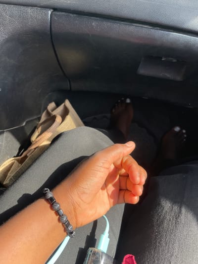 A human hand with a dark bead bracelet is resting on a person's lap in a car. The person is wearing black pants. The toes of the person's feet, painted white, are visible in the footwell of the car. A tan tote bag is near the person's leg. A blue phone charging cable is next to the person's hand. The location is unknown, there is no indication that it is in Dokwa, Nigeria.